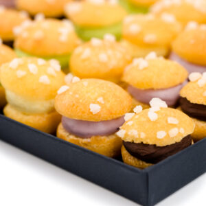 Vue rapprochée Les minis-tropézienne présentées sur un plateau rectangulaire en carton noir : vanille, chocolat, pistache, fruits rouges